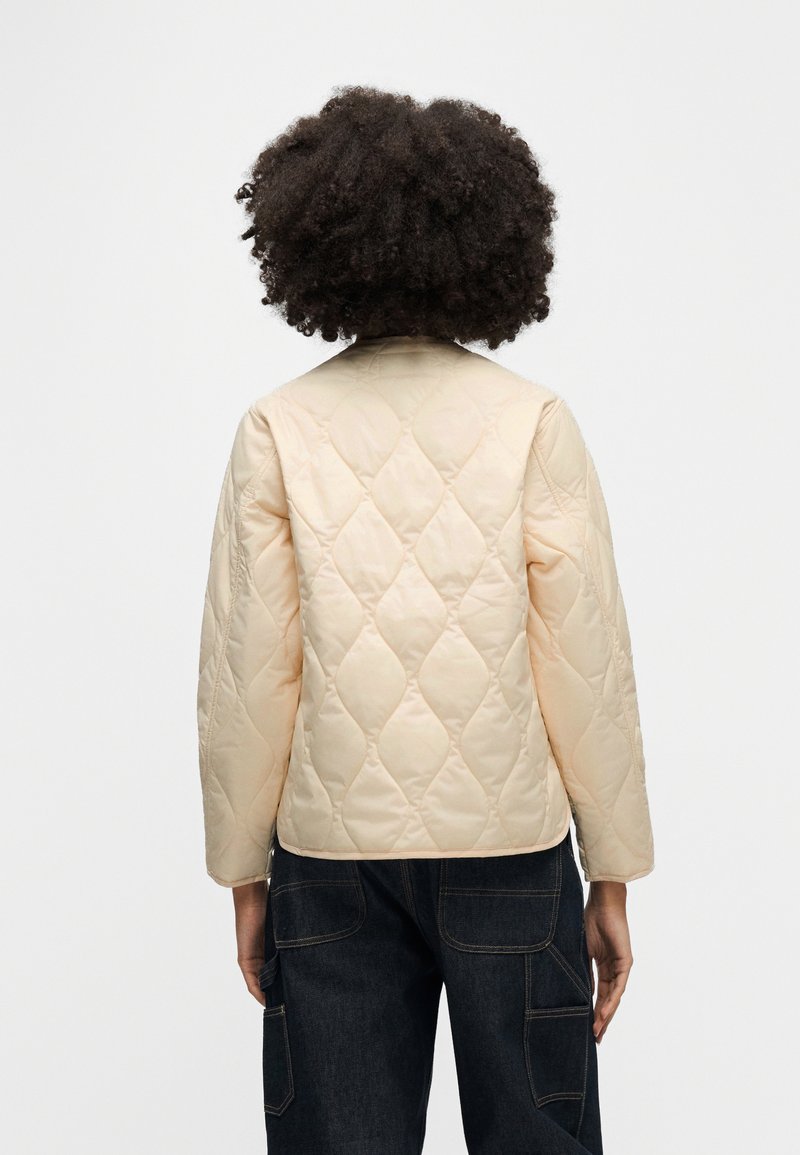 Persona con capelli ricci che indossa una giacca trapuntata beige chiaro e pantaloni scuri, vista da dietro su uno sfondo bianco.