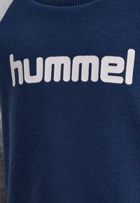 Granatowy sweter z wyściółką, z ribbingu przy szyi i dużym białym logo "hummel" na przodzie. Tekstura materiału jest miękka i gładka.
