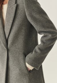 Abrigo de lana gris con un diseño entallado, que cuenta con solapa en pico, bolsillo lateral y puños acanalados visibles. Textura suave y forma estructurada.