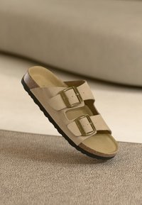 Beige Wildledersandale mit zwei verstellbaren goldenen Schnallen, gepolstertem Fußbett und einer strukturierten schwarzen Gummisohle. Einfaches, flaches Design.