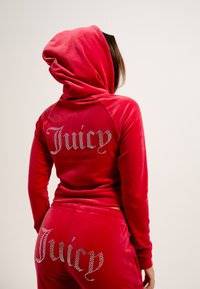 Tuta in velluto rosso con cappuccio; presenta la scritta 'Juicy' in strass sul retro del cappuccio e dei pantaloni; texture liscia, design aderente.