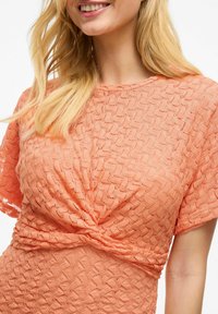 Haut de couleur pêche avec un motif texturé complexe, manches courtes et un détail noué à la taille. Tissu doux et léger.