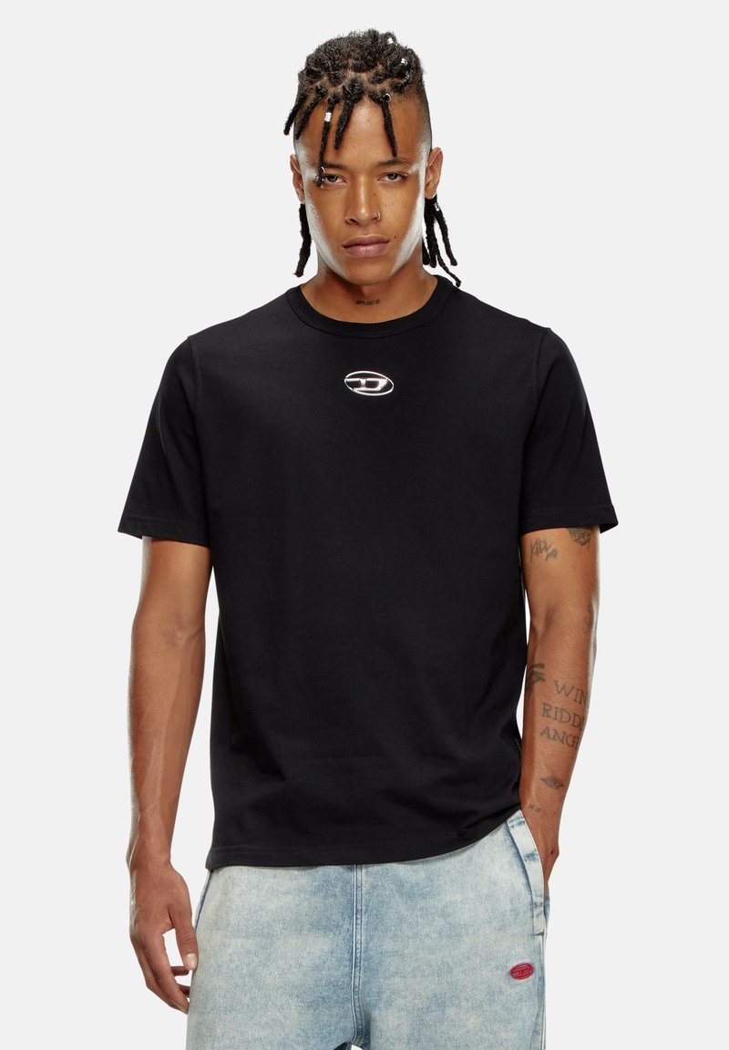 T-shirt en coton noir avec un col rond, des manches courtes et un petit logo métallique sur la poitrine. Modèle portant un short en denim bleu clair.