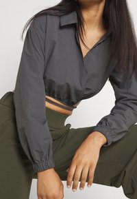 Zkrácená šedá košile s límcem, dlouhými nadýchanými rukávy a elastickými manžetami. Nositá přes kalhoty s vysokým pasem olivově zelené barvy. Hladká textura.