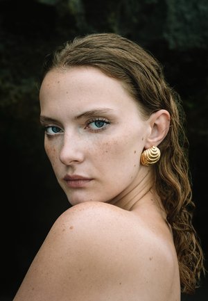Goldene Spiralohrringe, die am Ohr einer Person mit nassen braunen Haaren, strahlend blauen Augen und Sommersprossen präsentiert werden und eine glatte Hauttextur zeigen.