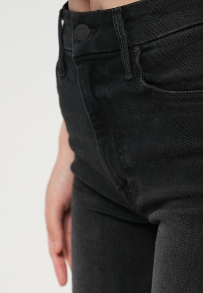 Close-up de uma pessoa a usar jeans de ganga pretos com um botão visível, passadores de cinto e parte de uma mão repousando ao lado do quadril.