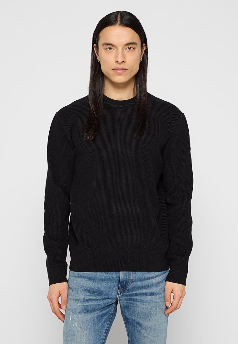 Maglione a coste nere con scollatura rotonda e maniche lunghe, abbinato a jeans chiari strappati. Design semplice e classico.