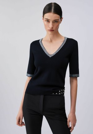 Donna che indossa una maglietta nera a scollo a V con rifiniture argento sulle maniche e sul collo, abbinata a pantaloni neri con dettagli borchiati.