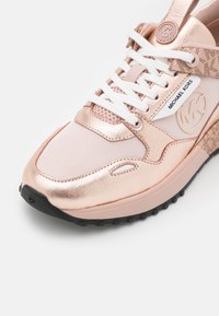 Baskets en or rose avec des lacets blancs, des panneaux en mesh, une semelle noire et un logo Michael Kors visible sur le côté et la languette.