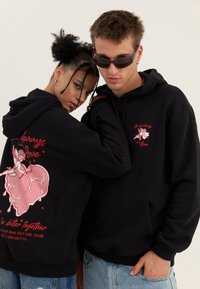 Two models wearing black hoodies with red cupid and heart graphics; one faces forward wearing sunglasses, the other shows back design.