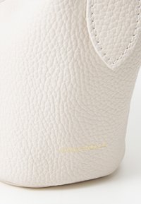Beige teksturert skinnveske med en buet form og en gull "Coccinelle"-logo på siden. Har glatte kanter og minimal maskinvare.