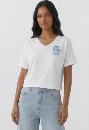 Frau mit langen schwarzen Haaren trägt ein weißes, bauchfreies T-Shirt mit V-Ausschnitt und blauem Motiv auf der linken Brust sowie hellblaue High-Waist-Jeans.