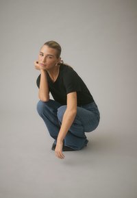 Melna apaļa kakla T-krekls, zilas augsta vidukļa džinsu bikses, tupus sēdoša poza, minimālas rotaslietas, gludi mati; smalkas tetovējumu zīmes uz apakšdelmiem.