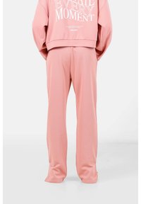 Sudadera rosa y pantalones a juego de tela suave. La sudadera tiene texto blanco, hombros caídos y un corte relajado; los pantalones son de corte recto.