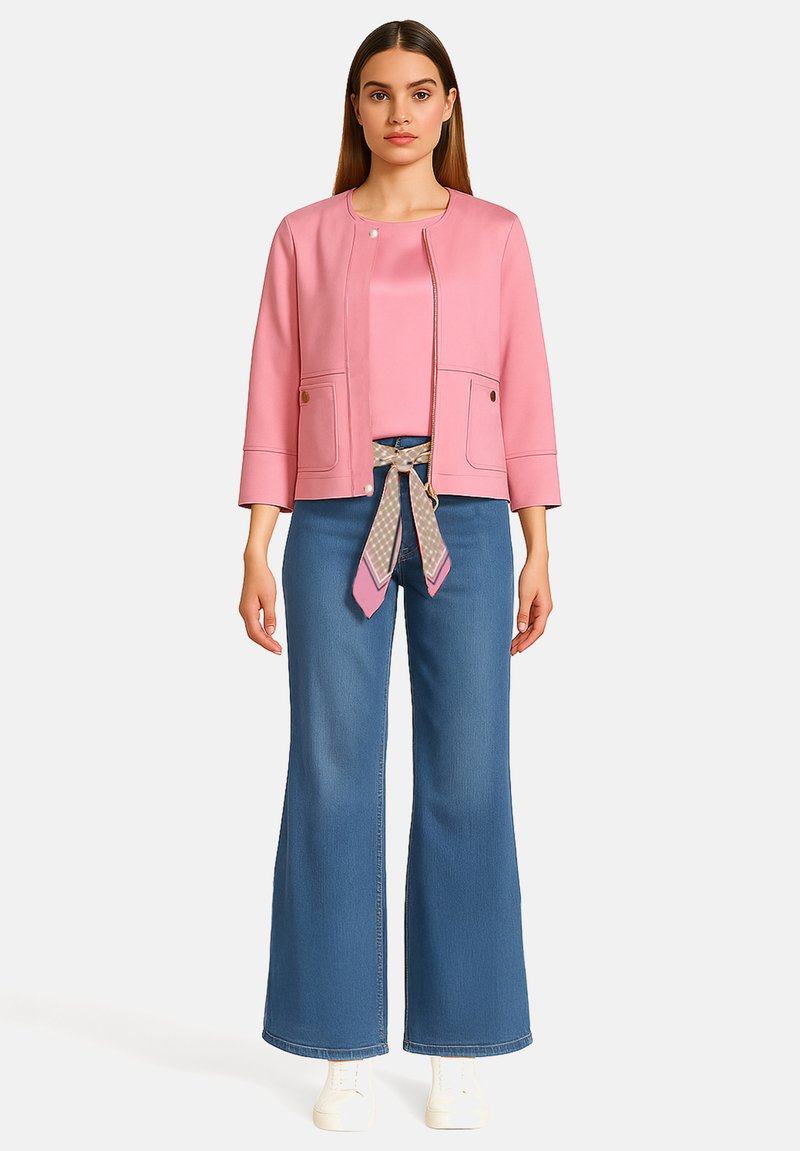 Veste rose à fermeture éclair avec deux poches et col rond ; associée à un haut rose et un jean bleu à jambes larges avec une ceinture à motifs. Baskets blanches.