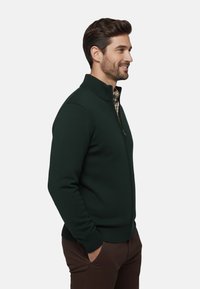 Sudadera de cremallera verde oscuro con textura acanalada, cuello alto y diseño ajustado, llevada sobre una camisa de cuadros, combinada con pantalones marrones.