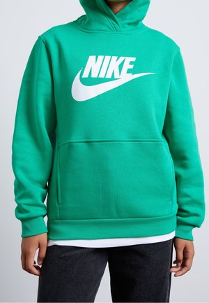 Osoba ubrana w turkusową bluzę z kapturem Nike z białym logo, ręce swobodnie opuszczone wzdłuż ciała, w połączeniu z ciemnymi spodniami i widoczną białą podkoszulką.