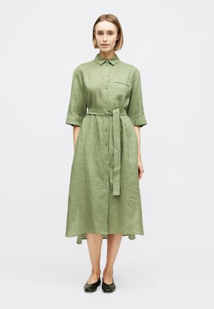 Woman wearing a light green button-up midi dress with a waist tie and black flats, standing against a plain white background.