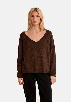 Donna bionda che indossa un maglione marrone largo a collo a V e pantaloni neri, in piedi su uno sfondo bianco uniforme.