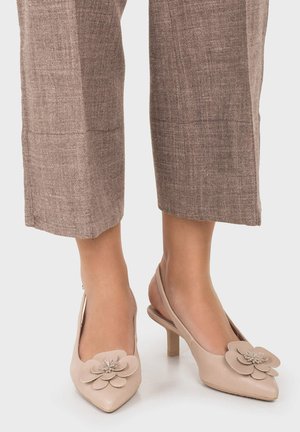 Zapatos de tacón beige con tiras traseras, puntera puntiaguda y grandes adornos florales, combinados con pantalones cortos marrones con textura.