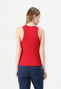 Rød ribbet tanktopp med høy hals og ermeløs design. Har en liten sløyfe bak. Kombinert med blå denimjeans.