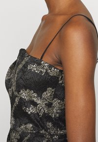 Robe noire en tissu texturé avec un motif floral doré. Fines bretelles noires et silhouette ajustée soulignant le torse.