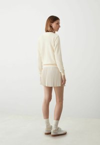 Crème-kleurige sweatshirt met geribde manchetten, gecombineerd met een geplooide beige minirok. Model draagt witte sneakers en bijpassende geribde sokken.