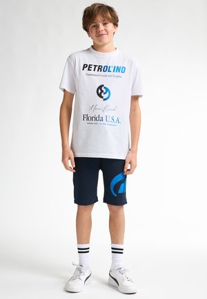Weißes T-Shirt mit schwarzem und blauem Text, kombiniert mit dunkelblauen Shorts mit einem blauen Logo. Dazu gehören gestreifte Socken und weiße Sneakers.
