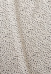 Tissu blanc avec de petites taches noires irrégulières réparties uniformément sur la surface, présentant de légers plis et une texture.