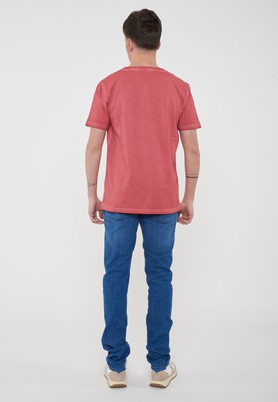 T-shirt rossa in cotone con maniche corte, colletto rotondo e vestibilità rilassata, abbinata a jeans blu e sneakers dai colori chiari.