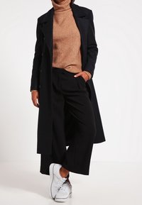 Abrigo negro largo, suéter de cuello alto marrón, pantalones anchos negros, zapatillas blancas. Texturas de tela suaves, corte a medida, diseño clásico.
