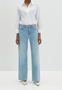 Světle modré široké džíny s vysokým pasem, které mají pět kapes, zapínání na knoflík a zip a jemné vyblednutí. V kombinaci s bílou košilí.