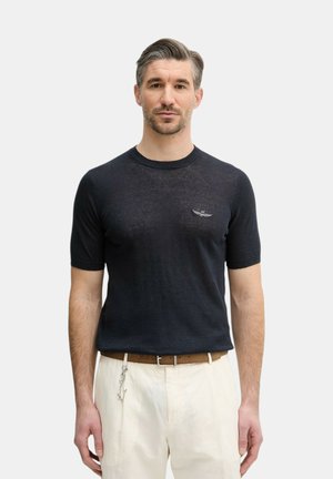 Uomo che indossa un maglione nero a maniche corte con un piccolo logo alato sul petto, pantaloni bianchi e una cintura marrone con un portachiavi a forma di stella.