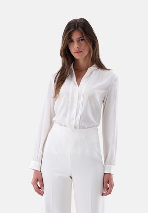 Frau mit langen braunen Haaren, die eine weiße, knopflose Langarmbluse und hoch taillierte weiße Hosen vor einem schlichten Hintergrund trägt.