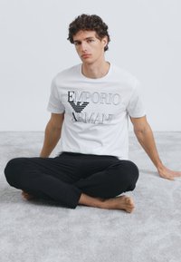 Balta kokvilnas T-krekls ar melnu "Emporio Armani" logotipu, kopā ar melnām biksēm. Modelis sēž uz gaiši pelēkas paklāja, basām kājām.