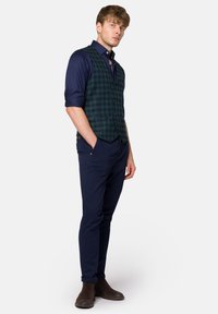 Zielona i granatowa kamizelka w kratę na ciemnoniebieskiej koszuli, w połączeniu z granatowymi spodniami i brązowymi botkami za kostkę. Gładki materiał, dopasowany krój.