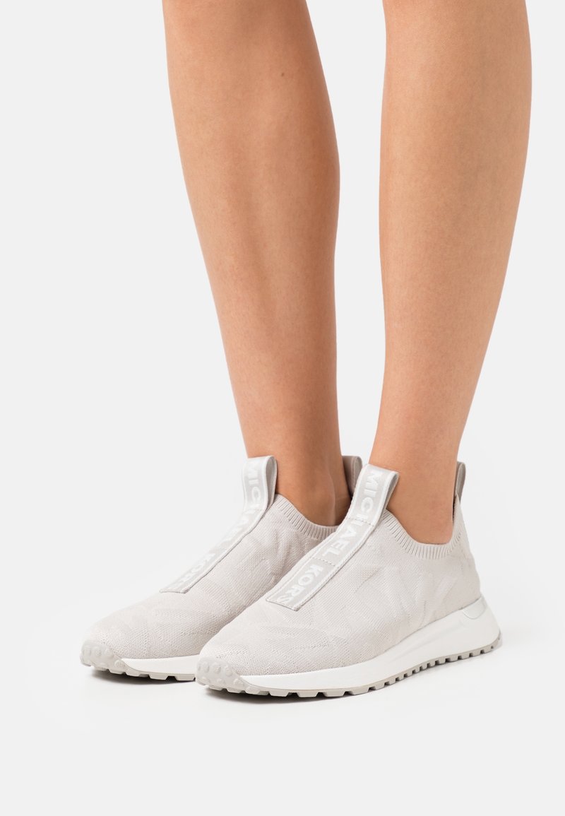 Personne portant des baskets à enfiler gris clair avec semelles blanches, dessus en maille élastique et languettes avec texte de la marque, debout sur un fond blanc.