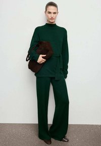 Suéter de punto verde con cuello alto y cintura ceñida, combinado con pantalones anchos a juego; sosteniendo un bolso de ante marrón oscuro.