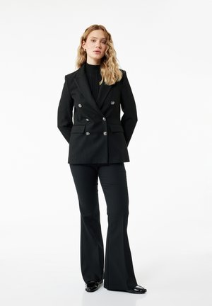 Frau steht und trägt einen schwarzen zweireihigen Blazer, schwarzen Rollkragenpullover, schwarze ausgestellte Hose und schwarze spitze Schuhe vor weißem Hintergrund.