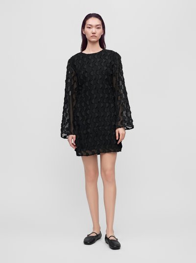 Un mannequin se tient devant un fond gris clair, vêtu d’une mini-robe noire à motifs et à manches longues, avec des chaussures plates noires.