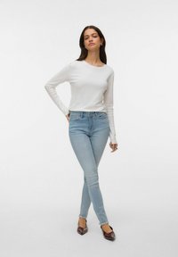 Balti ilgomis rankovėmis palaidinė derinama su šviesiai mėlynais liesais džinsais ir tamsiai rudos spalvos batais. Modelis atsipalaidavusio požiūrio stovi prieš paprastą foną.