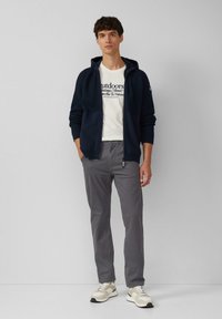 Pull à capuche marine avec texture côtelée, t-shirt graphique blanc, pantalon gris et baskets beiges avec des accents verts. Tenue décontractée avec des zips.