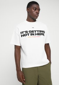 Jovem vestido com uma t-shirt branca com o texto em destaque "ESTÁ A FICAR QUENTE AQUI" e um texto menor abaixo, acompanhada por calças cargo verde-oliva.