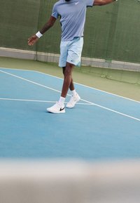 Vaaleansininen urheilupaita ja kuvioidut shortsit, joissa on Nike-logo; valkoiset tennarit mustilla yksityiskohdilla sinisellä tenniskentällä.