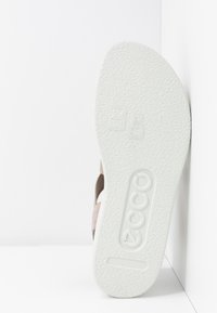 Semelle texturée blanche d'une sandale affichant la taille 38 et le logo de la marque "ecco" en relief, positionnée verticalement contre un mur blanc.