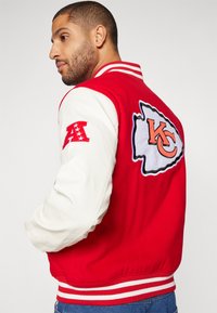 Rdeč šolski jopič z belimi usnjenimi rokavi, ki ima na hrbtu velik logotip KC, rebrast ovratnik in črtast rob.