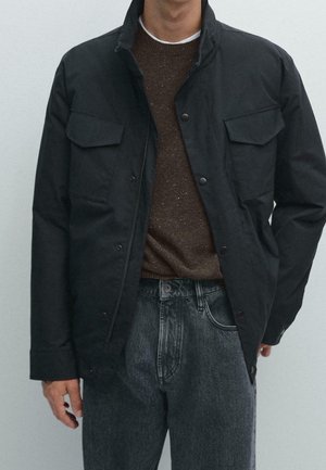 Homme portant une veste noire boutonnée sur un pull marron et un jean gris foncé, debout devant un fond clair uni.