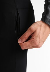 Pantalon de sport noir pour homme en tissu extensible, doté d'une poche zippée sur le côté. La texture est lisse avec une coupe ajustée et des coutures renforcées.