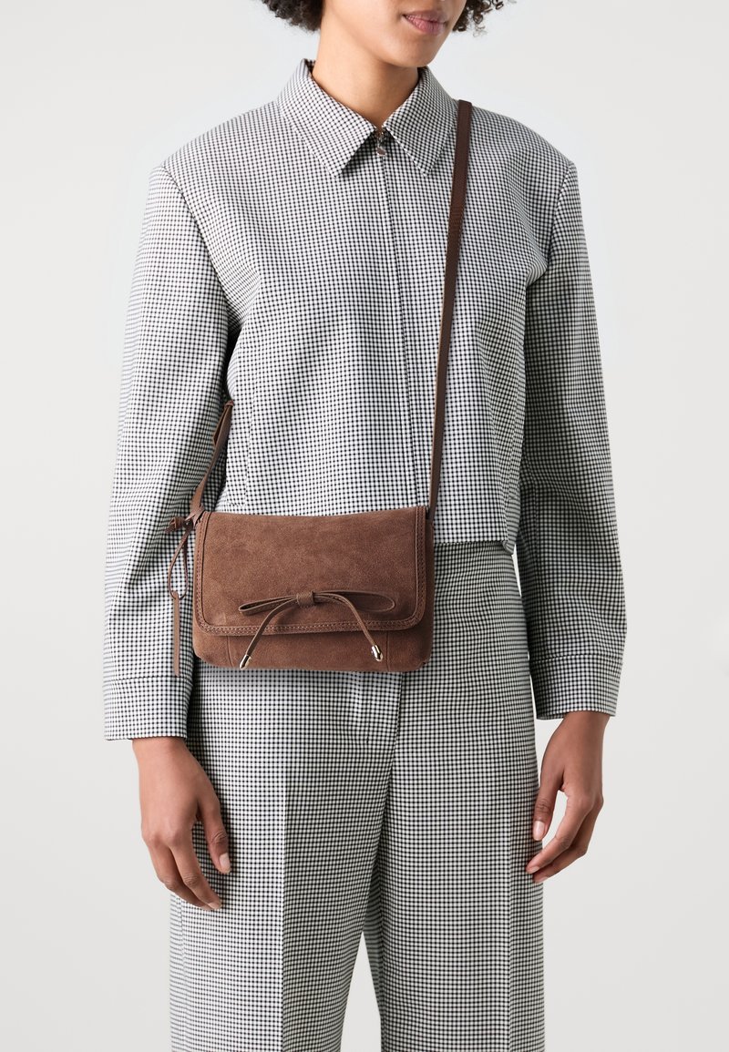 Sac en suède marron en bandoulière avec une forme plate, un détail noué à l'avant et des accents argentés. Porté sur une tenue à carreaux noir et blanc.