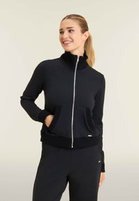 Chaqueta negra con cremallera hecha de una tela suave, con un alto cuello acanalado, dobladillo y puños acanalados, y dos bolsillos delanteros.